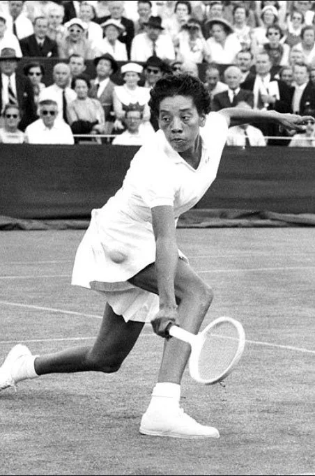 Black and white photo of a tennis player in a white outfit and sneakers, preparing to hit a shot on a tennis court with an audience watching in the background.