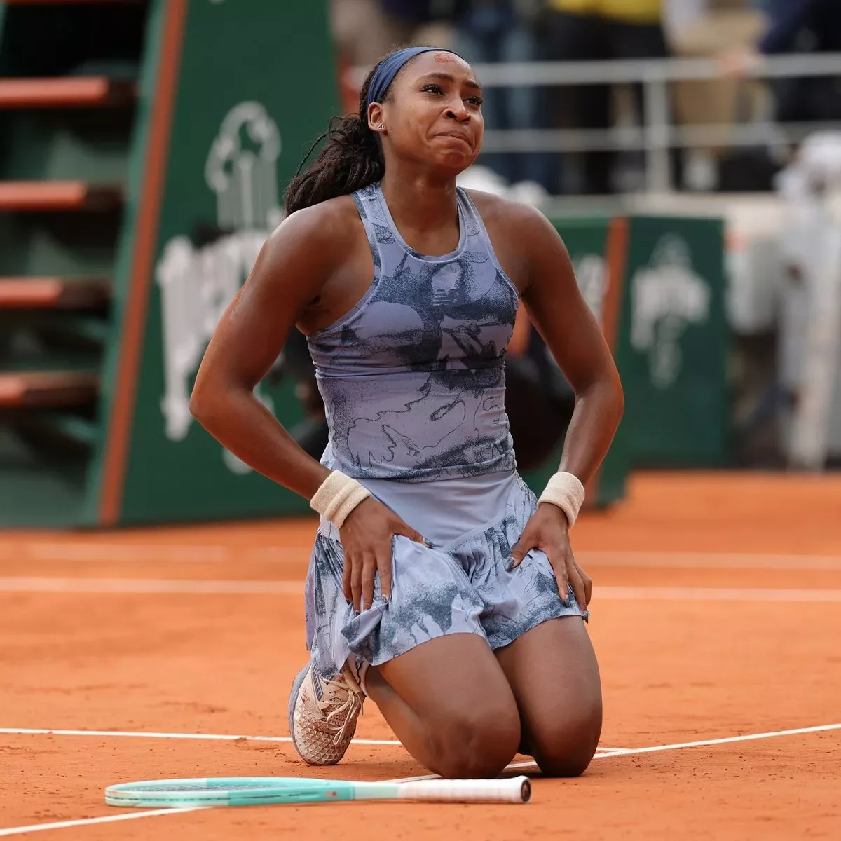 A female tennis player kneeling on a clay court, crying with her hands on her thighs, holding a tennis racket on the ground beside her.