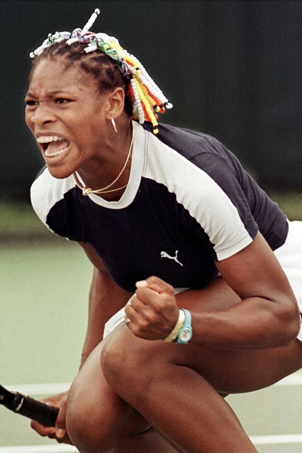 A woman with braided hair decorated with colorful beads, wearing a black and white Puma sports shirt, appears to be celebrating on a tennis court, with a determined and joyful expression.