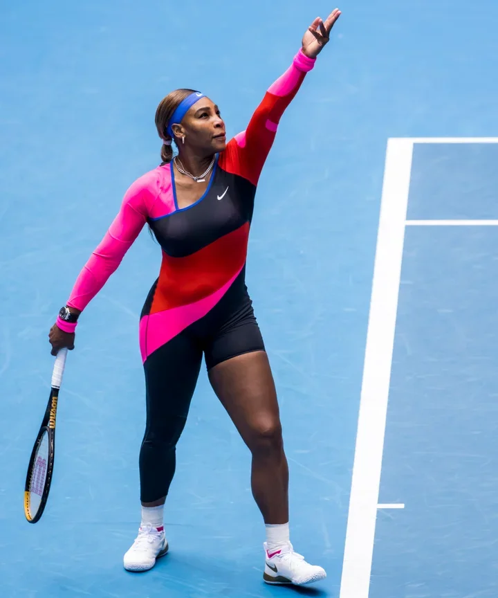 Tennis player on a blue court, holding a tennis racket, serving or striking a ball, dressed in colorful sportswear with a headband.