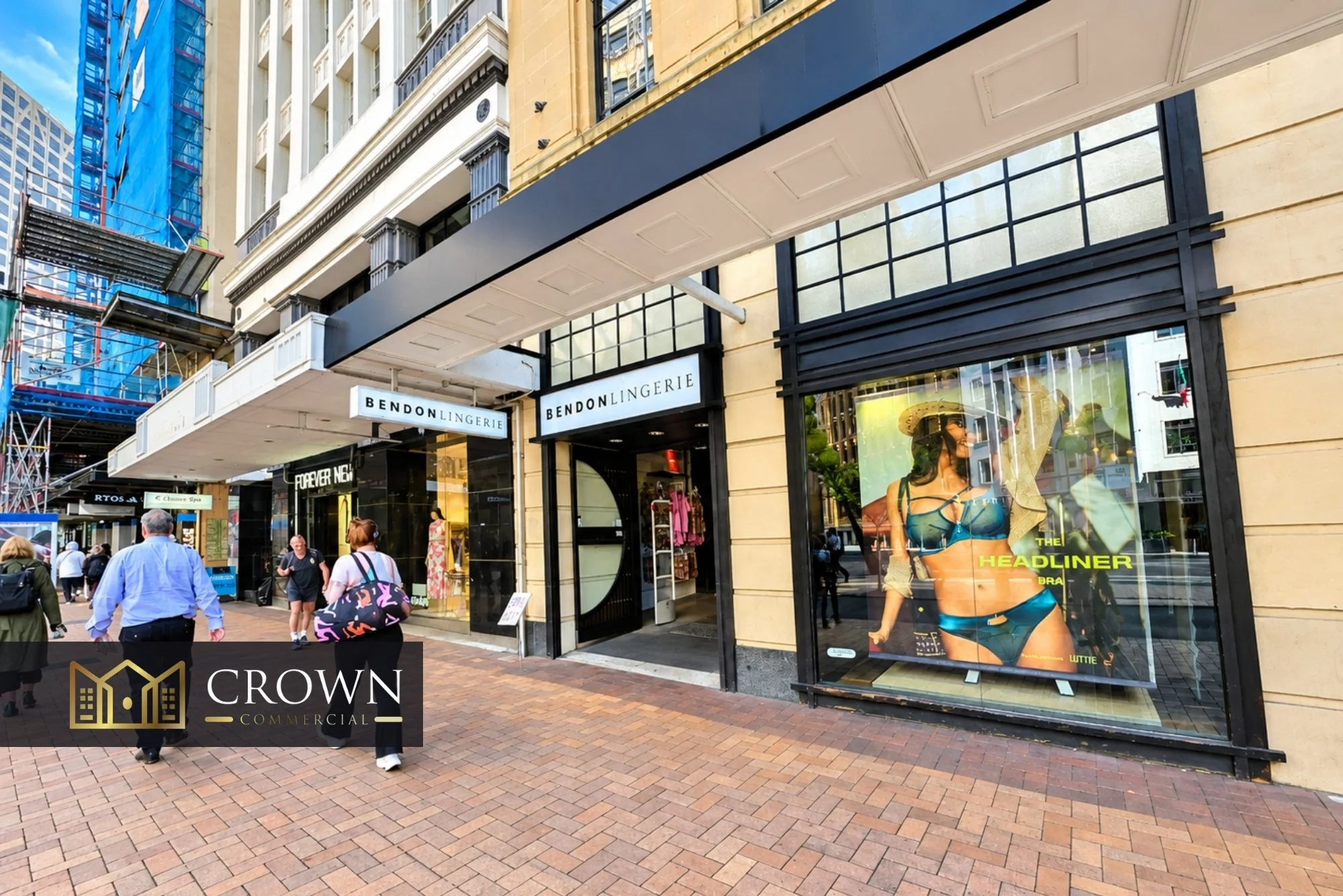 A busy shopping street with stores including Bendon Lingerie, with several pedestrians walking on the brick-paved sidewalk. A large advertisement displays a woman in lingerie in the store window.