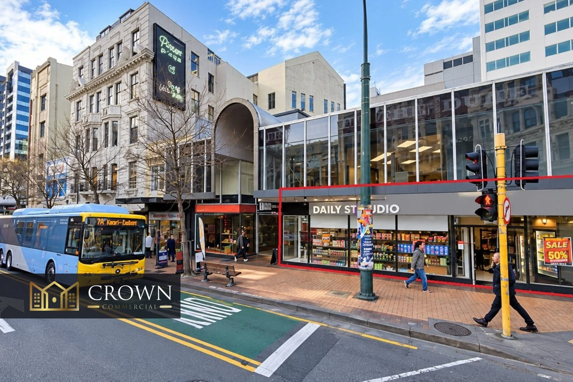 City street scene with a blue bus, pedestrians, a store named Daily Studio with a large window, a traffic light, leafless trees, and tall buildings under a partly cloudy sky.