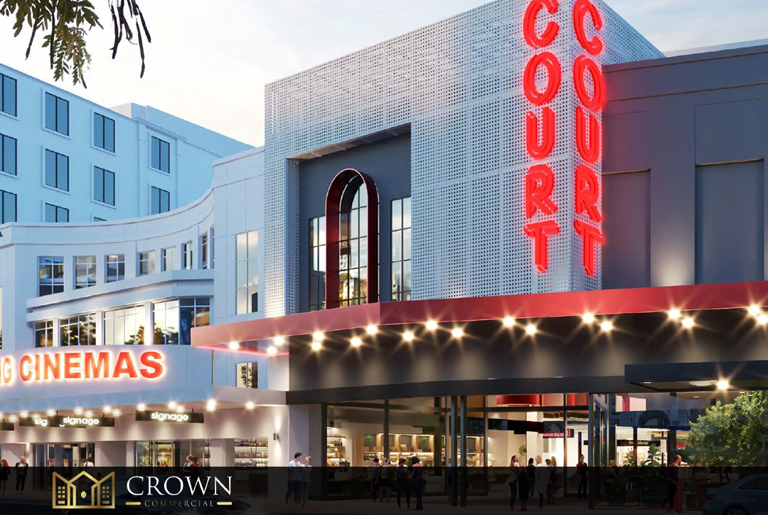 Exterior view of a modern movie theater with illuminated signage, including a vertical red sign reading 'COURT' and a marquee with bright lights, glowing in the evening.