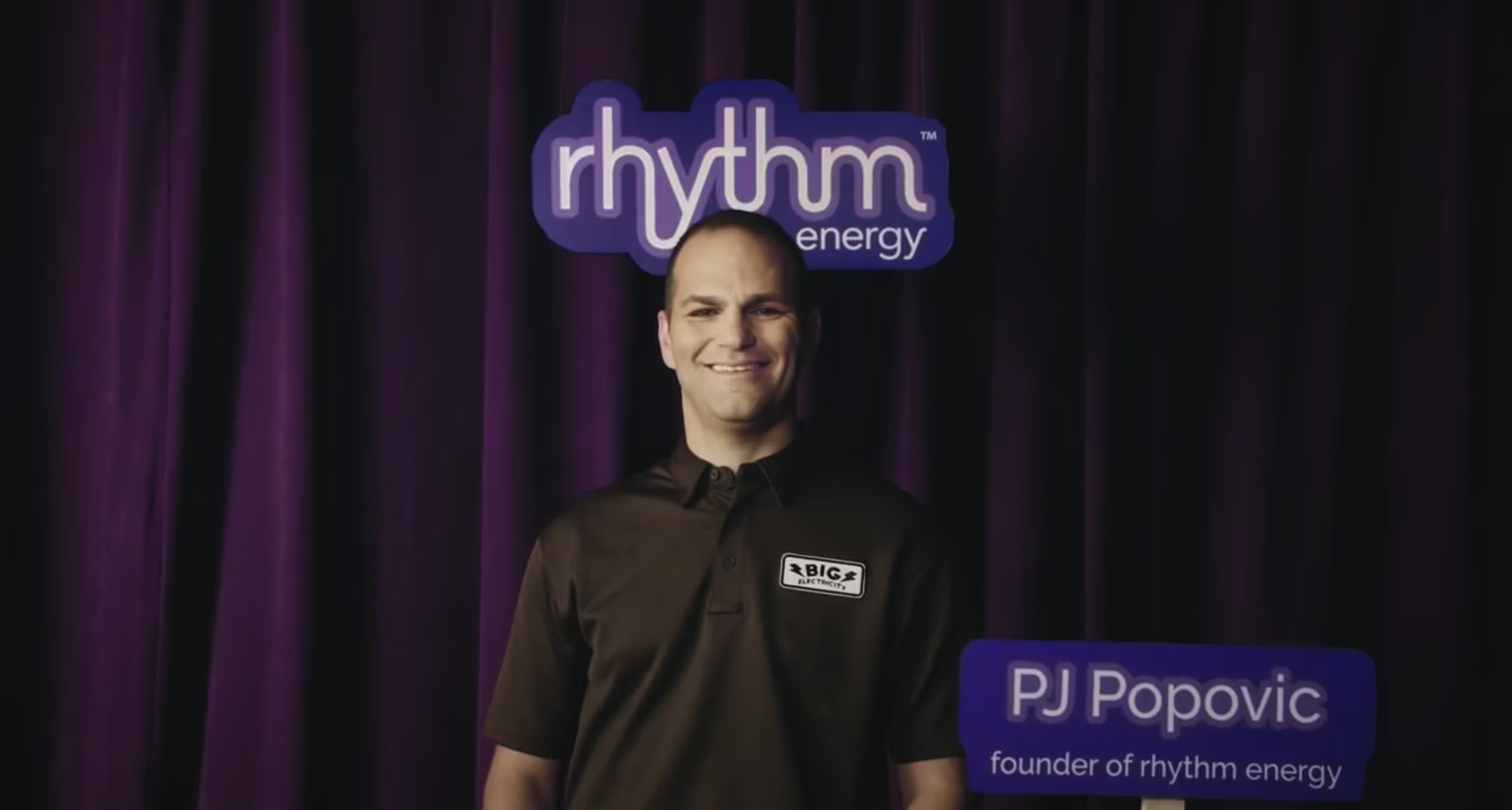 Man smiling in front of purple curtain with 'rhythm energy' sign, wearing a black shirt with 'BIG ELECTRICITY' logo, and a caption identifying him as PJ Popovic, founder of rhythm energy.