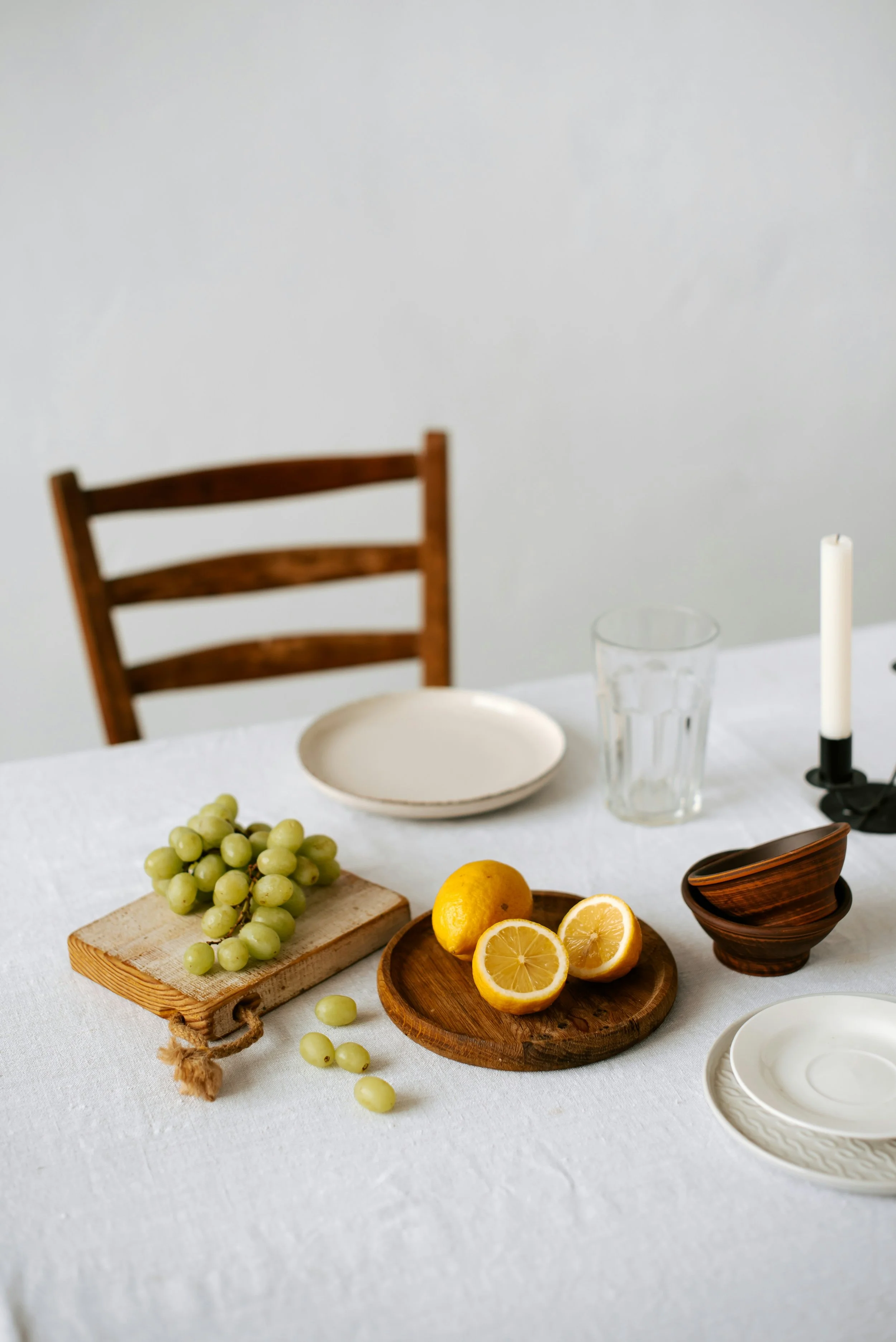 A table set with a bunch of green grapes, a lemon cut in half, a half of lemon on a wooden plate, two empty white plates, glasses, a candlestick with a white candle, and stacked bowls.