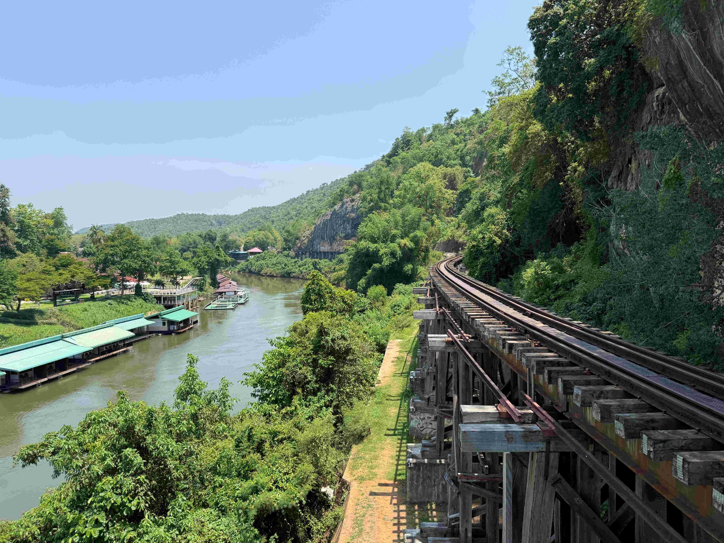 Train tracks running along a mountainside with dense green foliage and hills covered with trees extending into the distance, while a river flows beneath on the left side with floating raft structures all under a bright blue sky.