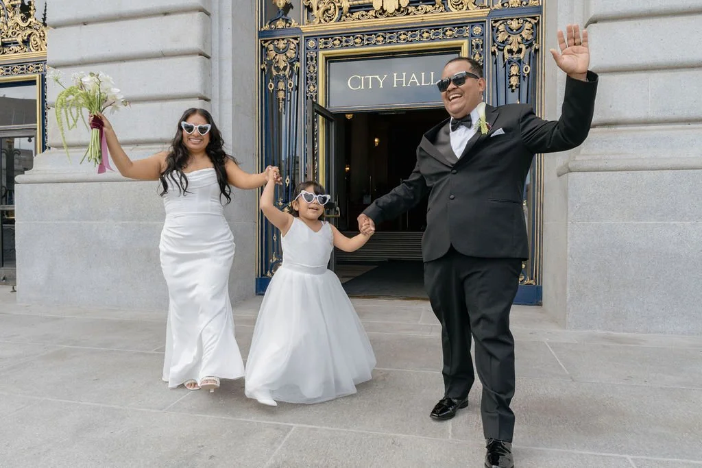 Just-married couple celebrating their San Francisco City Hall wedding while walking hand in hand with their daughter, all smiling in chic wedding attire and sunglasses.