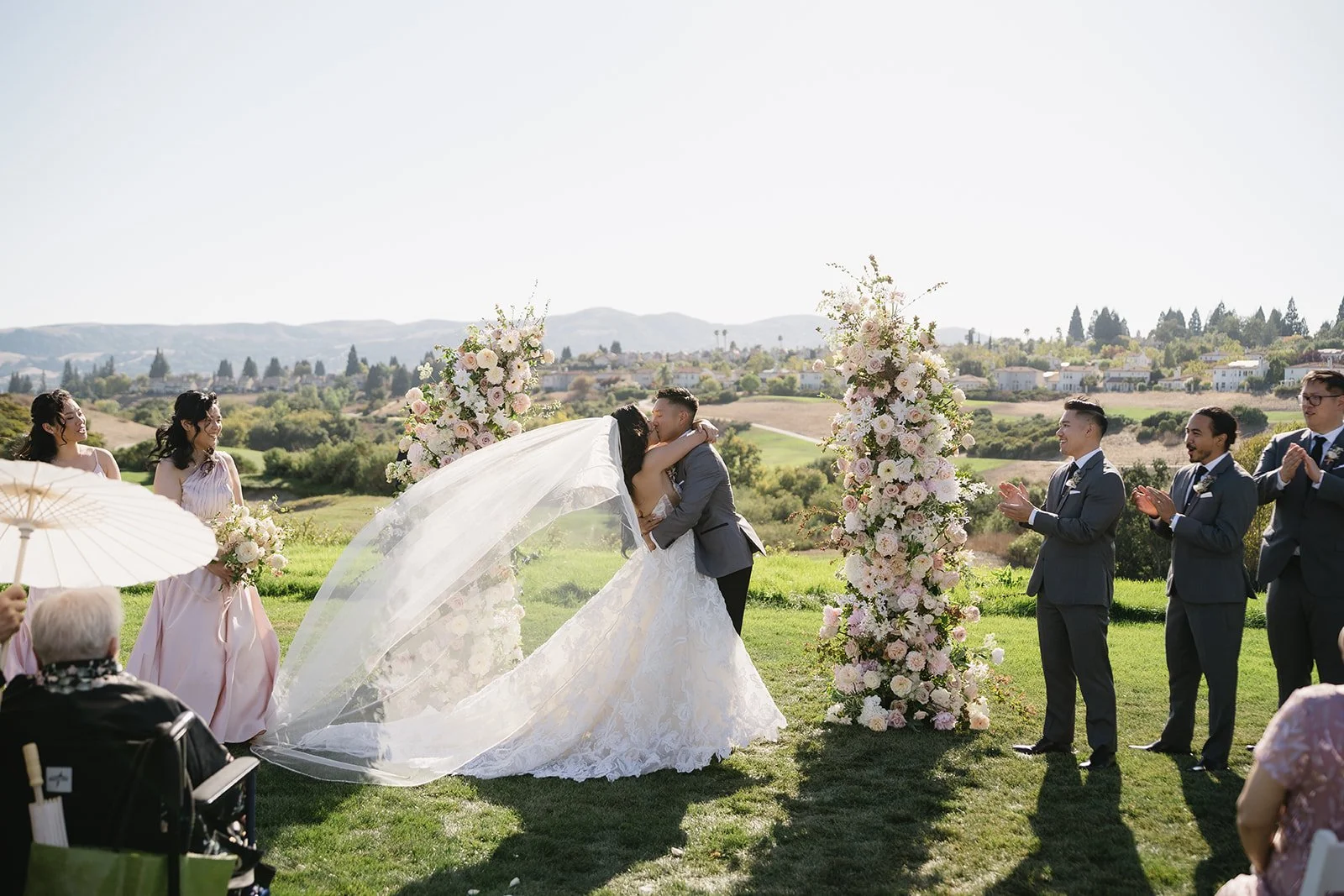 Bay Area outdoor wedding ceremony with bride and groom sharing their first kiss beneath blush and ivory floral pillars, flowing cathedral veil caught in the breeze, bridesmaids in blush gowns and groomsmen in gray suits, overlooking rolling hills and