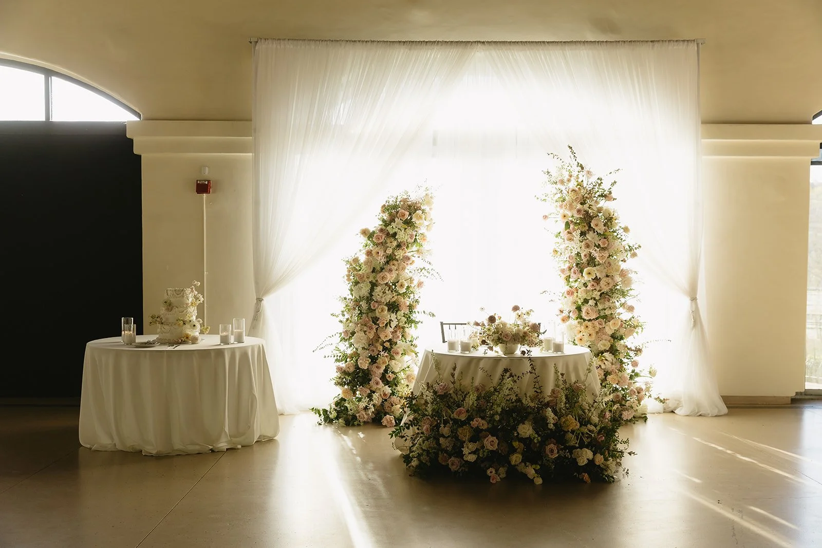 Elegant Bay Area wedding reception sweetheart table with lush blush and ivory floral columns, romantic white draped backdrop, cascading garden style greenery, candlelight details, and classic tiered wedding cake display in a modern indoor venue.