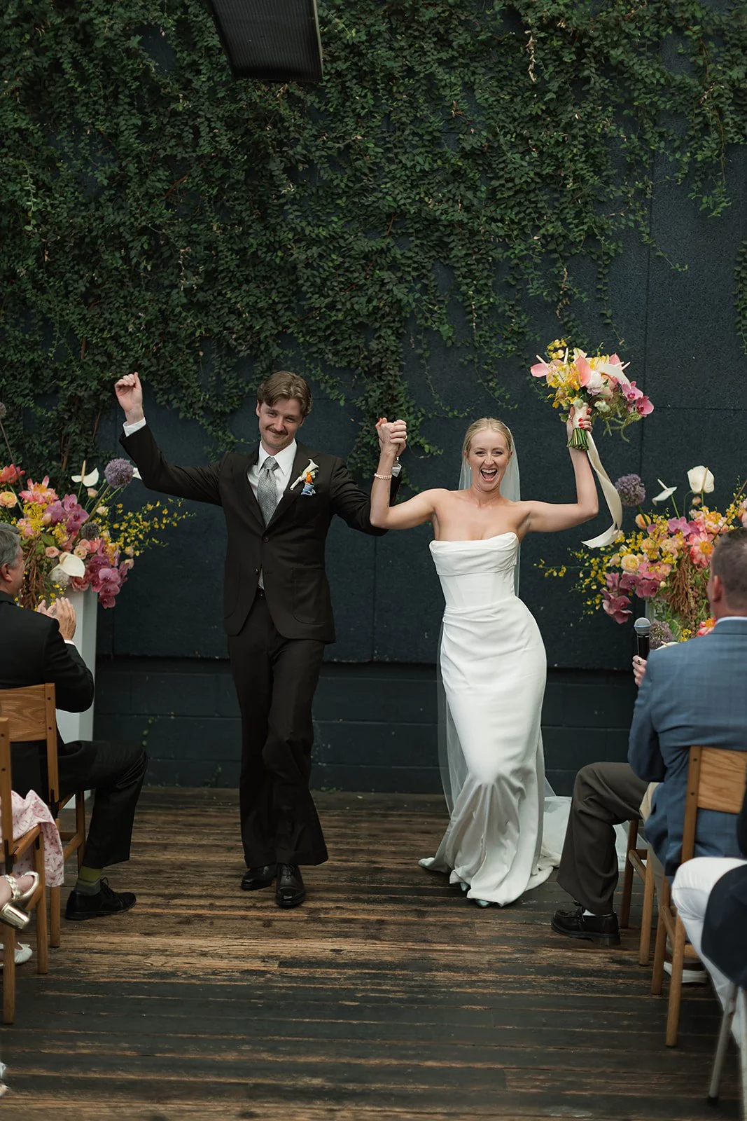 Berkeley wedding ceremony at Comal Restaurant with bride and groom celebrating down the aisle, modern white gown and black suit, vibrant colorful florals, ivy covered industrial backdrop, and intimate indoor reception setting in the Bay Area