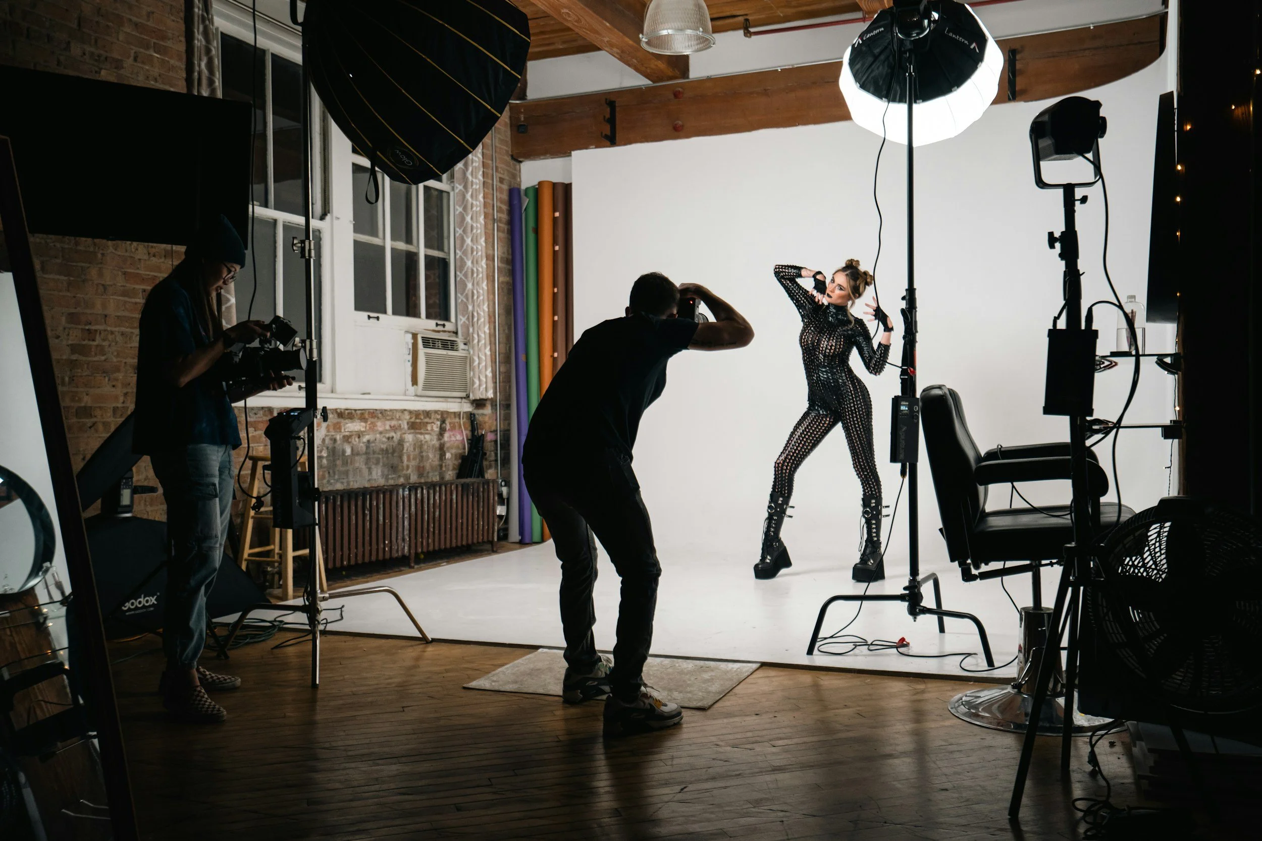 A photo studio with a model posing in front of a white backdrop, surrounded by lighting equipment and photographers. The model is wearing a black, see-through, studded outfit and has her hair styled in space buns.