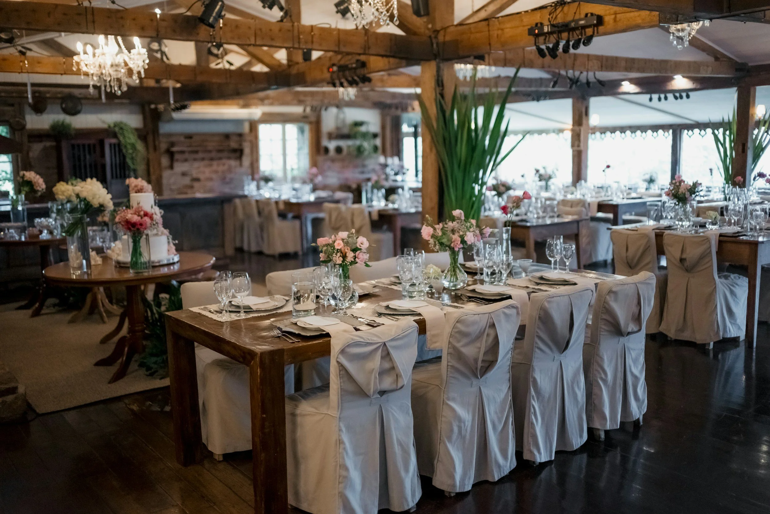 Wedding reception setup with round table decorated with flowers, surrounded by chairs with white covers, in a rustic venue with wooden beams and chandeliers.