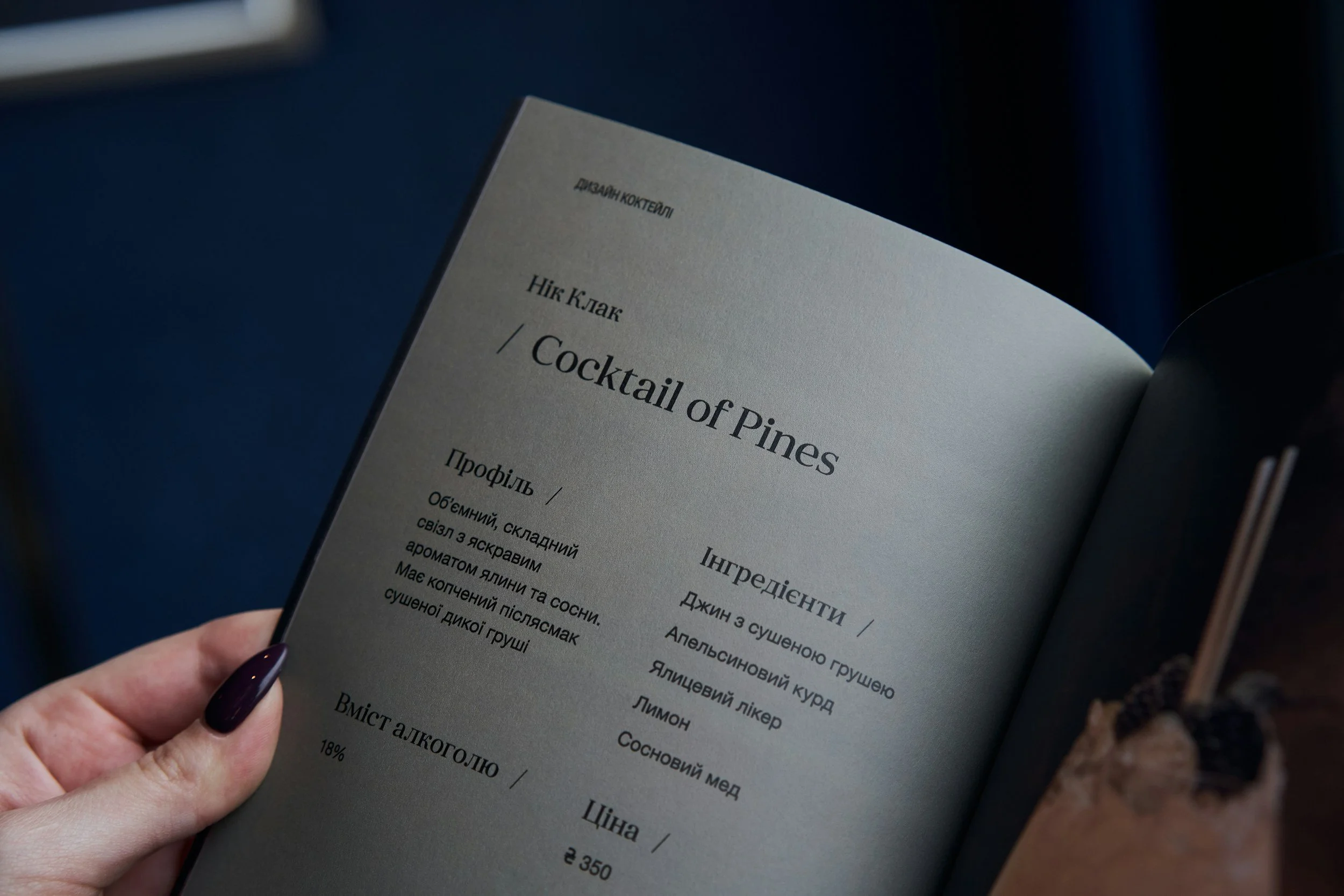 A person holding an open menu showing a cocktail recipe called 'Cocktail of Pines' written in English and another language, with some ingredients and instructions visible.