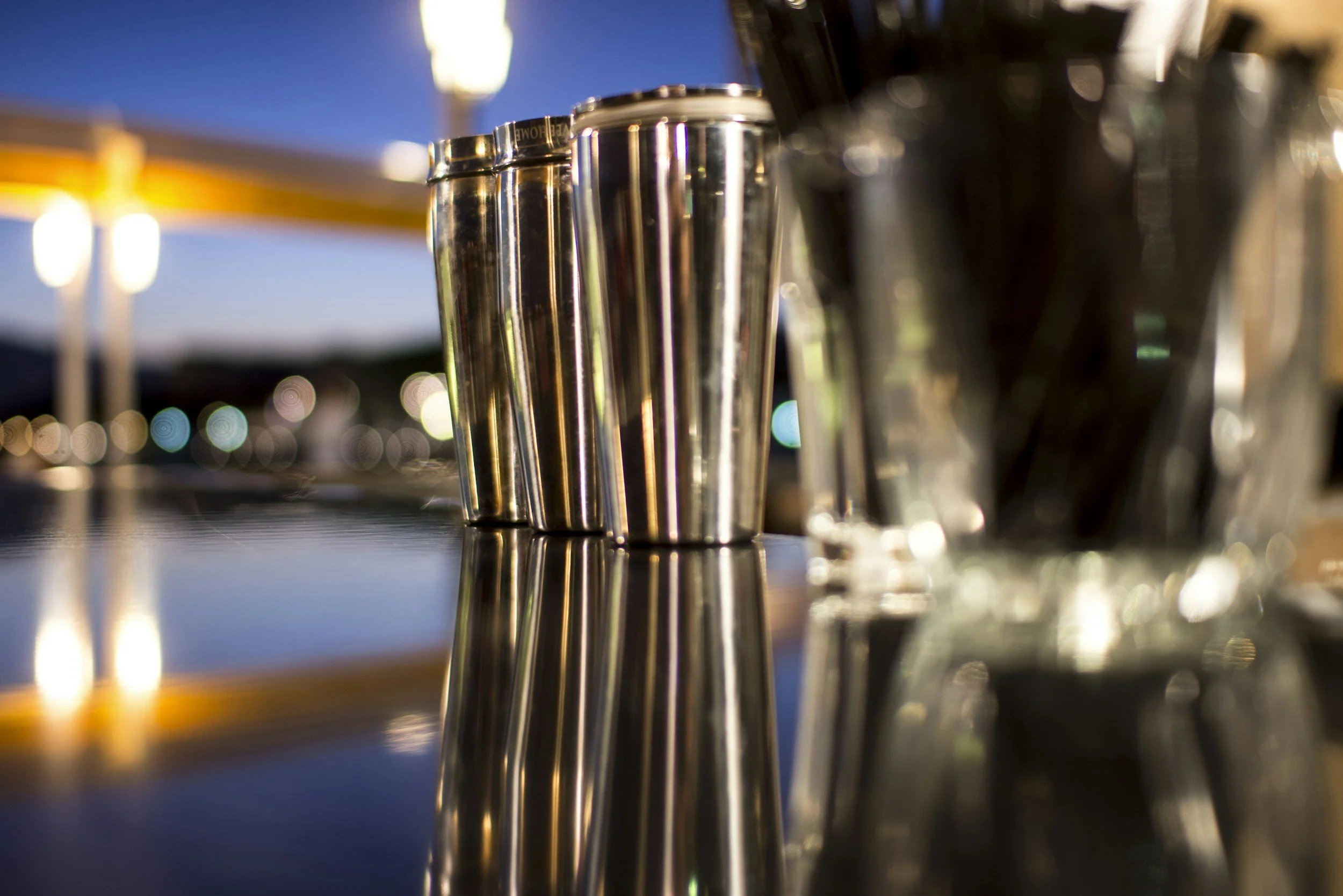 Close-up of shiny stainless steel cocktail shakers on a reflective surface, with a blurred background of a sunset sky.