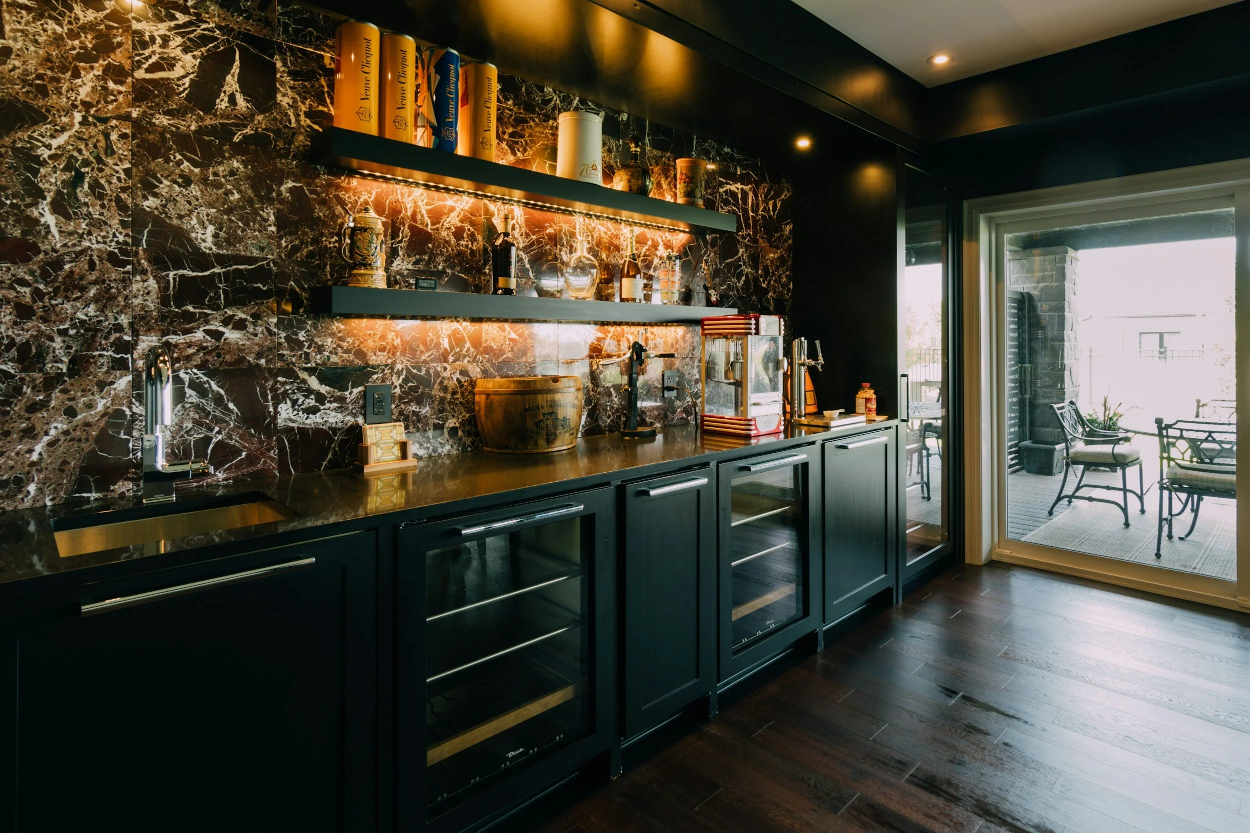 A modern kitchen featuring dark cabinetry, a black marble backsplash with white veining, and open shelves displaying various decorative items and bottles. There is a sliding glass door leading to an outdoor patio with chairs and a table, allowing natural light to brighten the space.