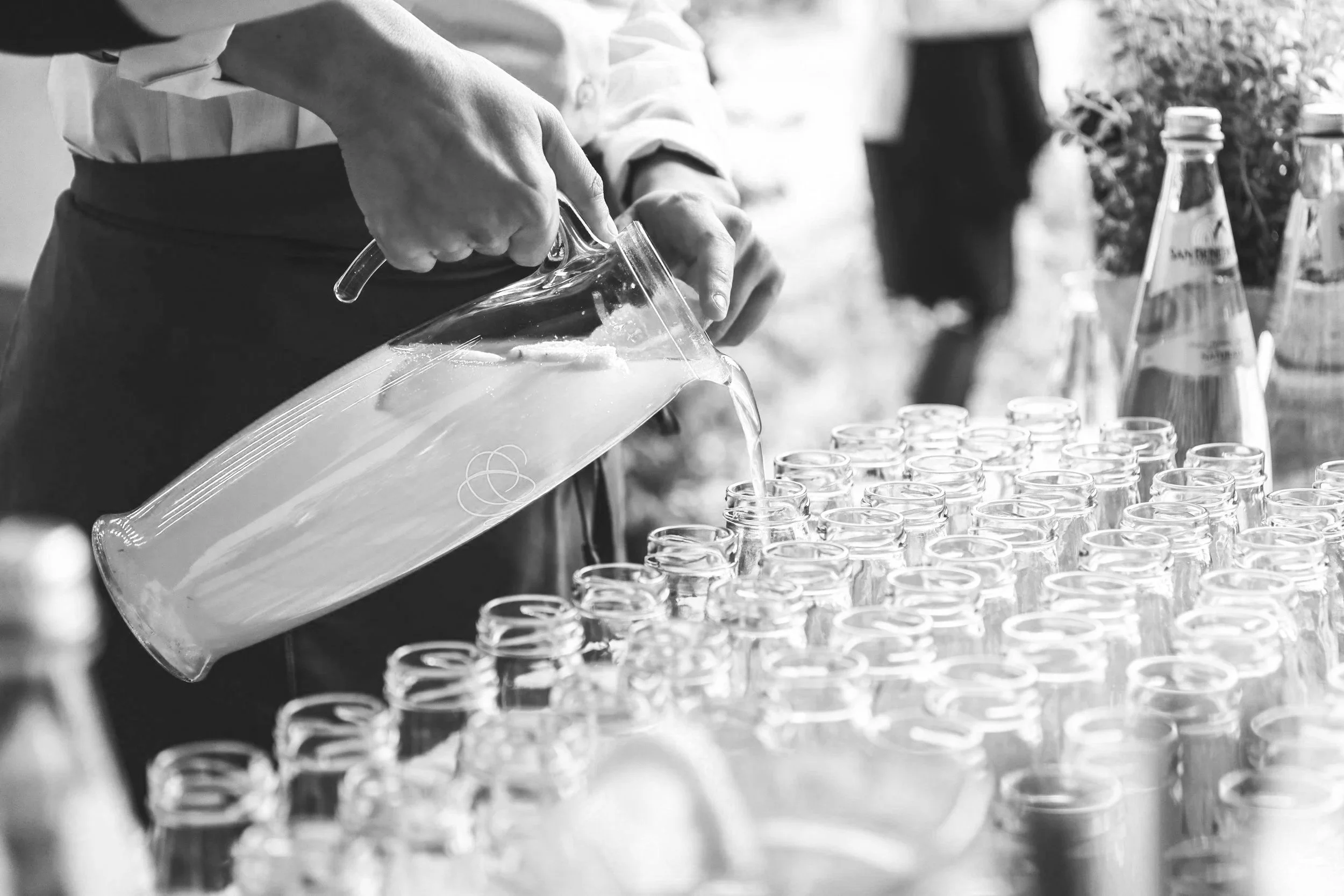 A person pouring liquid into small glass cups arranged on a table, with bottles of water in the background. The image is in black and white.