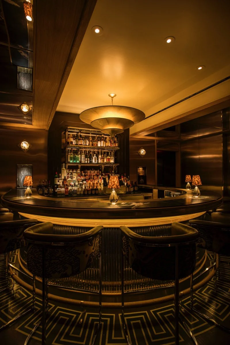 A luxurious bar with a circular counter, shelves of liquor bottles behind it, small lamps on the bar, and comfortable bar stools, all decorated in warm, dim lighting.
