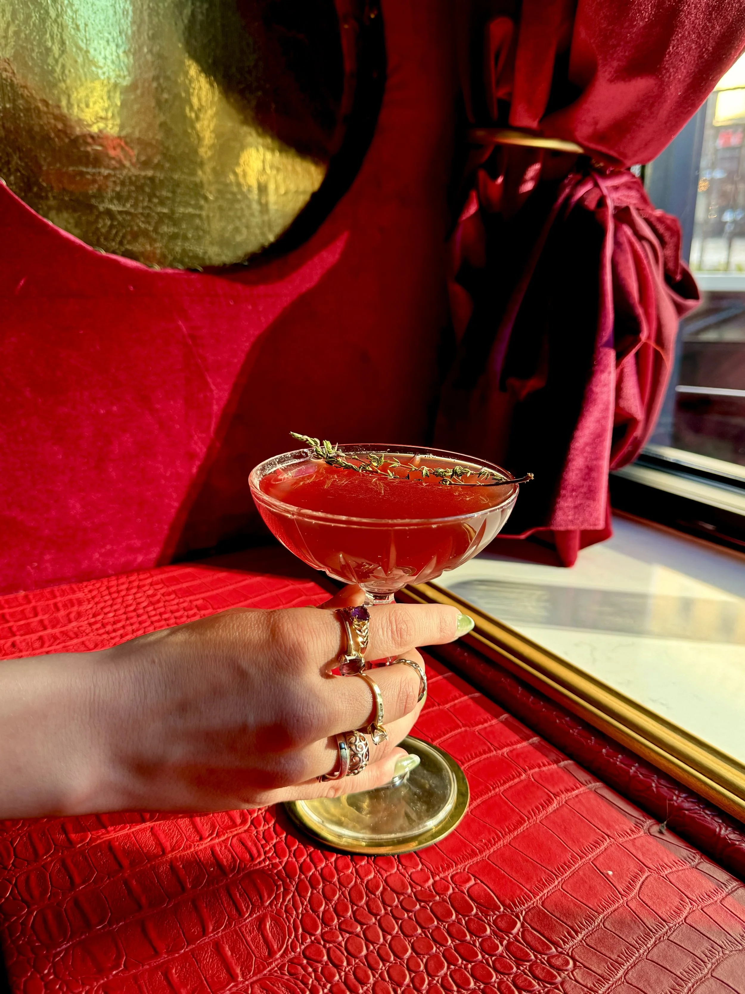 A hand holding a cocktail glass with a sprig of thyme on top, resting on a red crocodile textured surface. Someone wearing rings has their hand on the surface near the glass, with a person in a red jacket in the background.