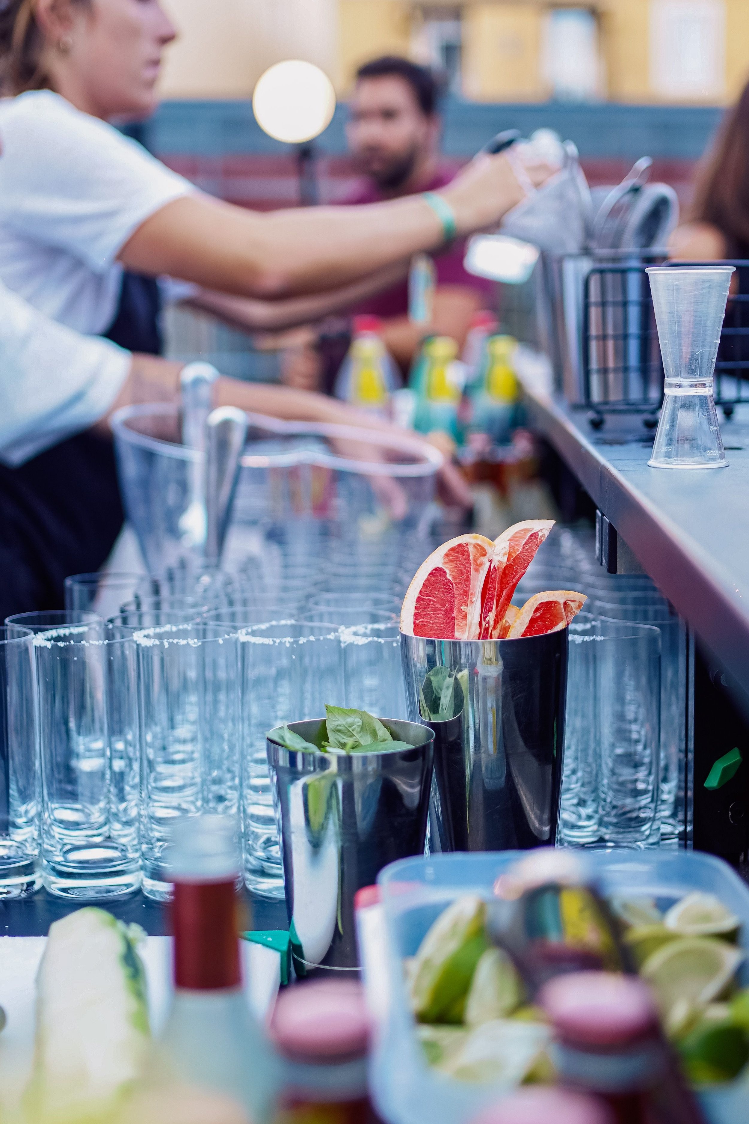 A bartender preparing drinks with garnishes like grapefruit slices and lime slices on a bar counter. Several empty glasses and bar tools are visible.