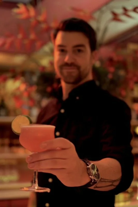 A man with dark hair and a beard holding a pink cocktail garnished with lemon, in a dimly lit, colorful bar or restaurant.