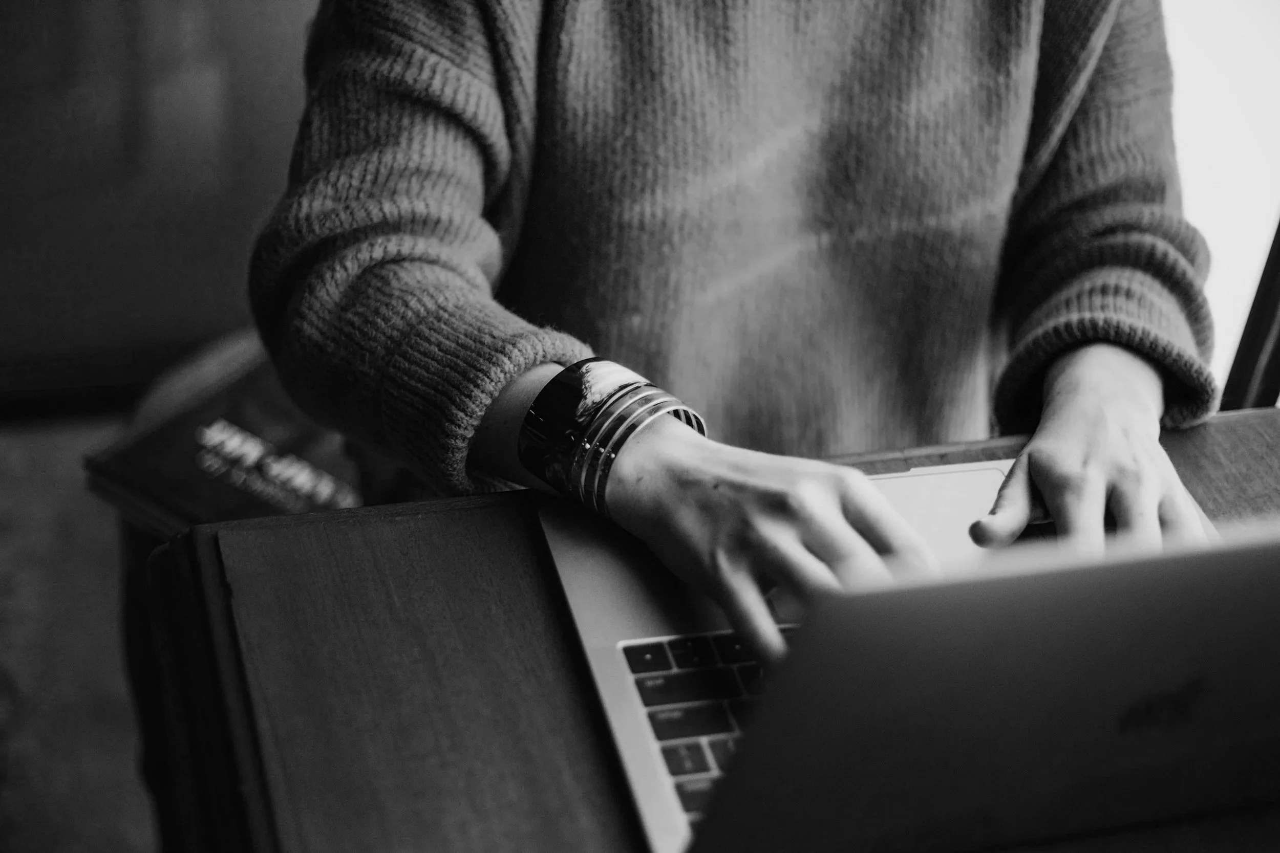 Black and white photograph of a person wearing a ribbed sweater, leaning over a table with their hands resting on it, in front of a laptop. The person's face is not visible.