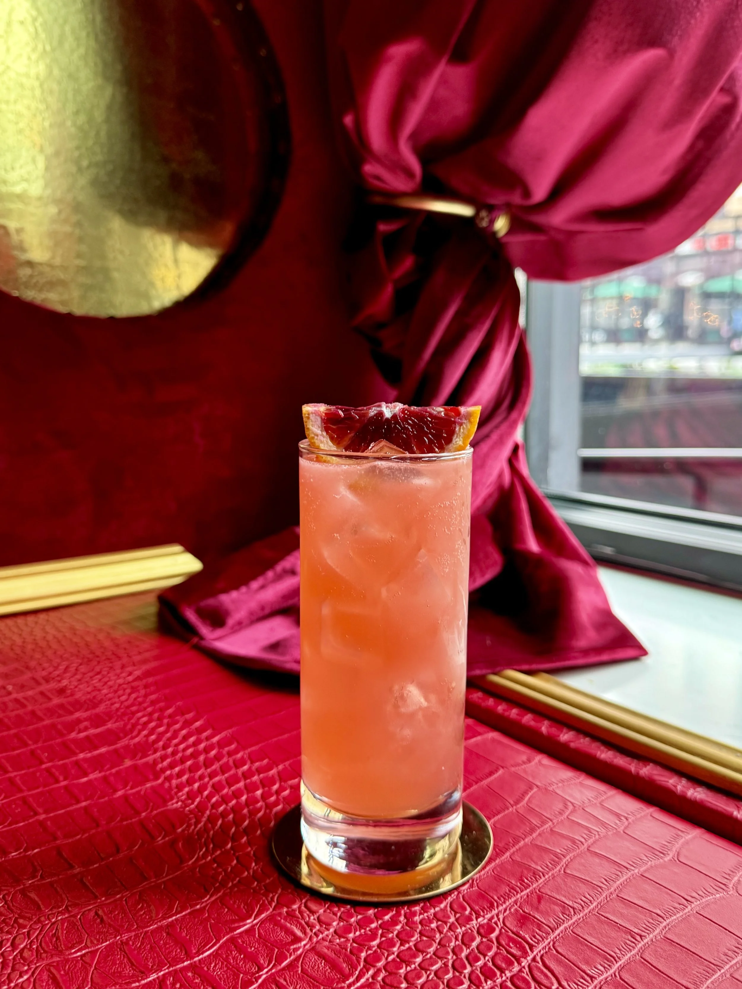 A tall glass of pink grapefruit cocktail garnished with a slice of grapefruit, set on a red textured table with a pink velvet cloth and golden chopsticks in the background.