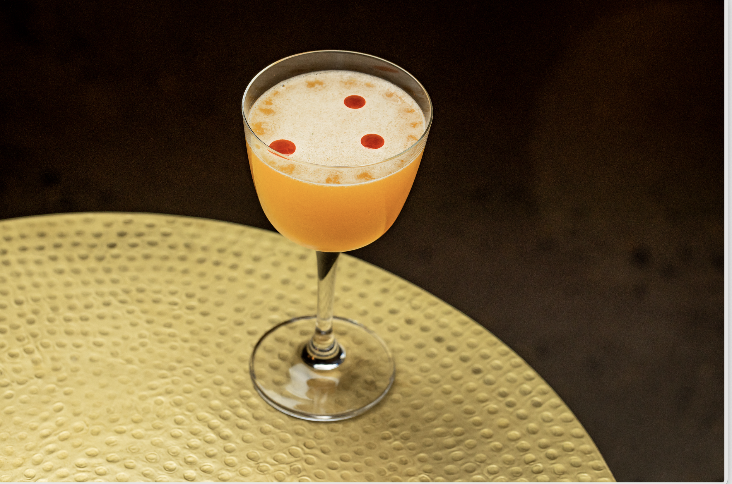 A glass of orange-colored beverage with foam on top, garnished with three small red drops, placed on a yellow rubber mat.