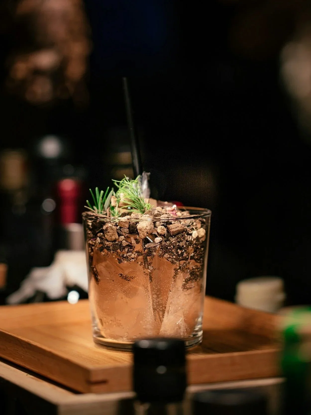 Close-up of a cocktail in a clear glass with a layered design, topped with decorative herbs and chocolate-like toppings, sitting on a wooden tray in a dimly lit setting.
