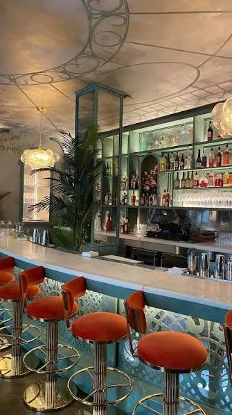 A stylish bar with orange bar stools around a curved white counter, a tall glass display cabinet, shelves with bottles and glasses, modern lighting fixtures, and decorative plants in a trendy interior.