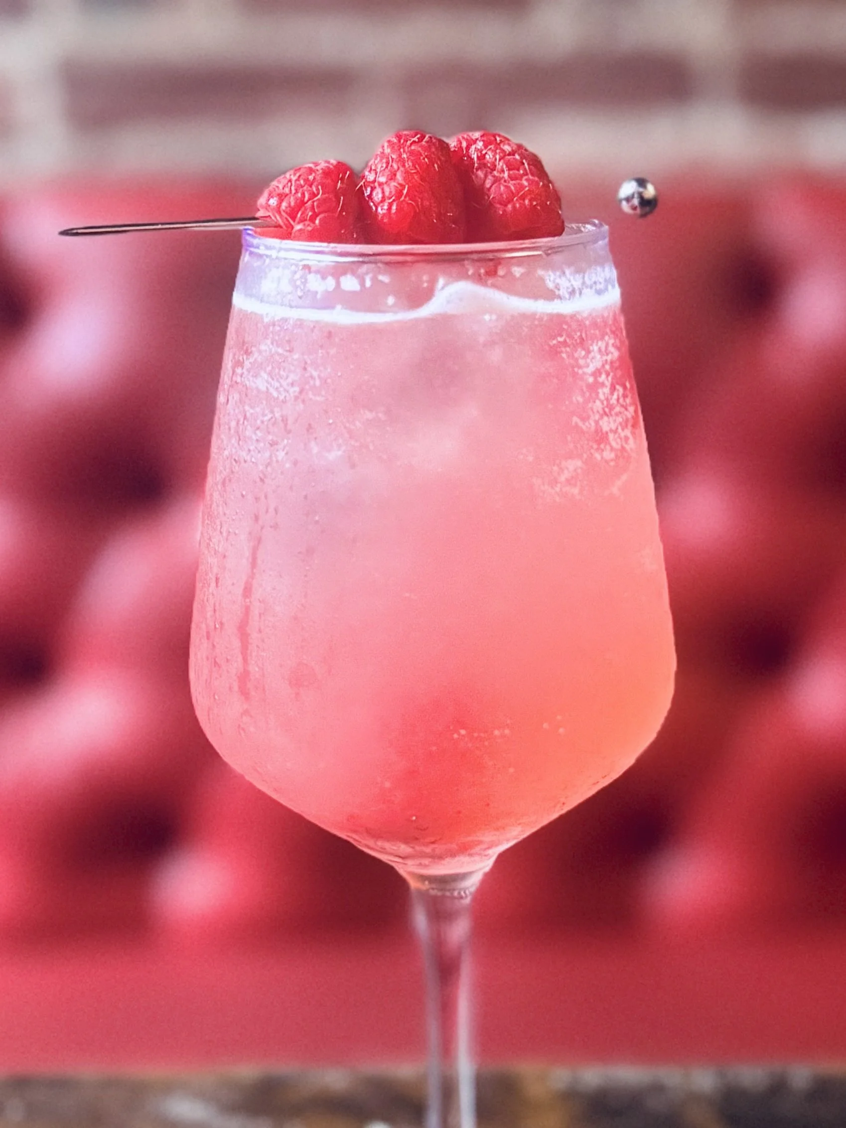 A close-up of a glass filled with a pink beverage, garnished with fresh raspberries, and a metal skewer resting on top, with a blurred background of more raspberries.