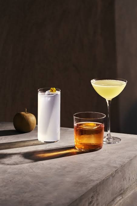 A still life of three cocktails on a gray stone surface, with an apple in the background. The drinks include a clear glass with a white cocktail garnished with a small flower, a short glass with an amber-colored drink garnished with an orange slice, and a tall martini glass with a pale yellow drink.