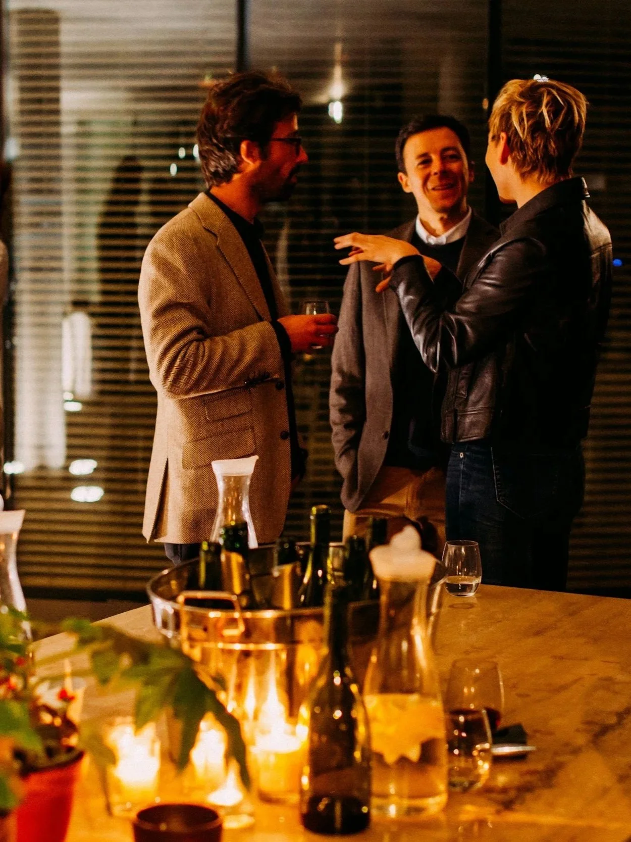 Three people are engaged in conversation at a social gathering in a warmly lit room, with drinks in their hands, a table with bottles and glasses, and candlelight in the foreground.