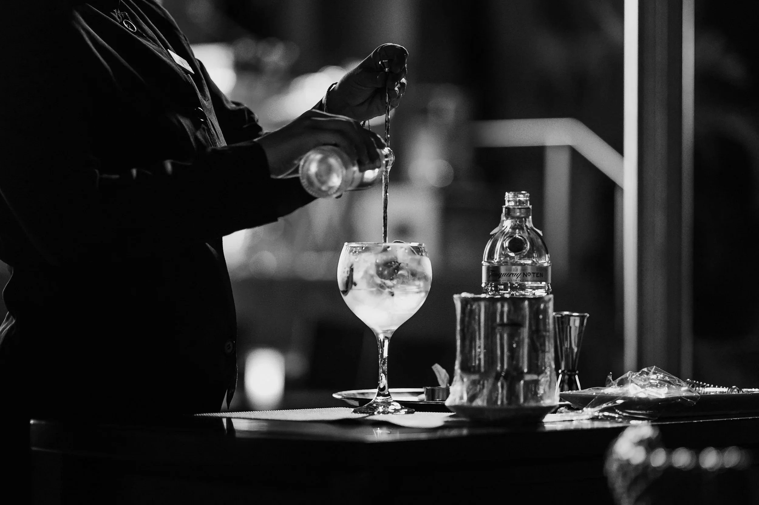 A bartender pouring a drink into a glass at a bar counter, with bottles of alcohol and glasses on the table in a black-and-white photo.