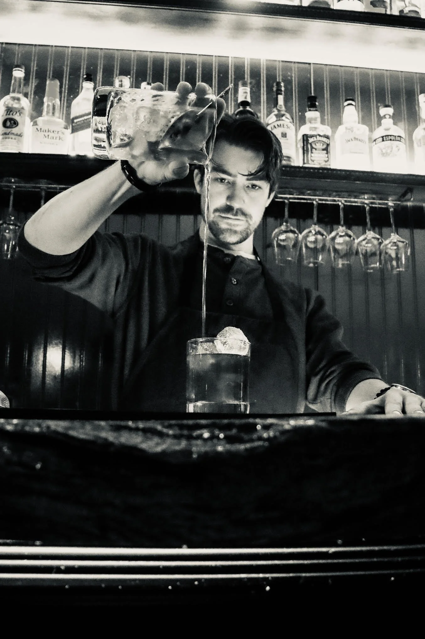 A man with dark hair and a beard pouring a drink into a glass with ice, standing in front of a bar with bottles of alcohol on shelves and wine glasses hanging upside down.