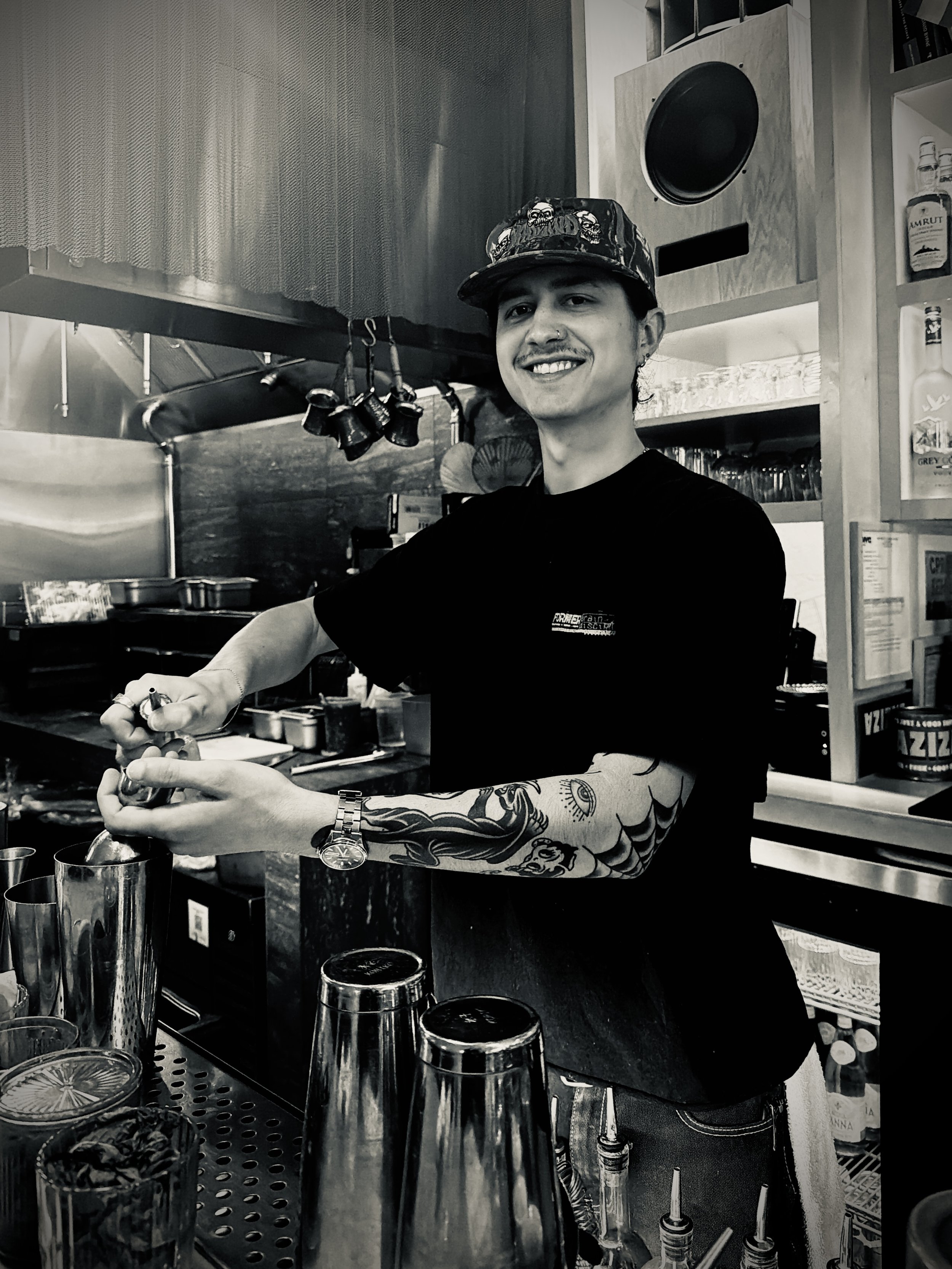 A smiling young person wearing a cap with skulls, a black T-shirt, and tattoos on their arm, working behind a bar or counter with various cans, glasses, and bar tools, in a bar or cafe setting.