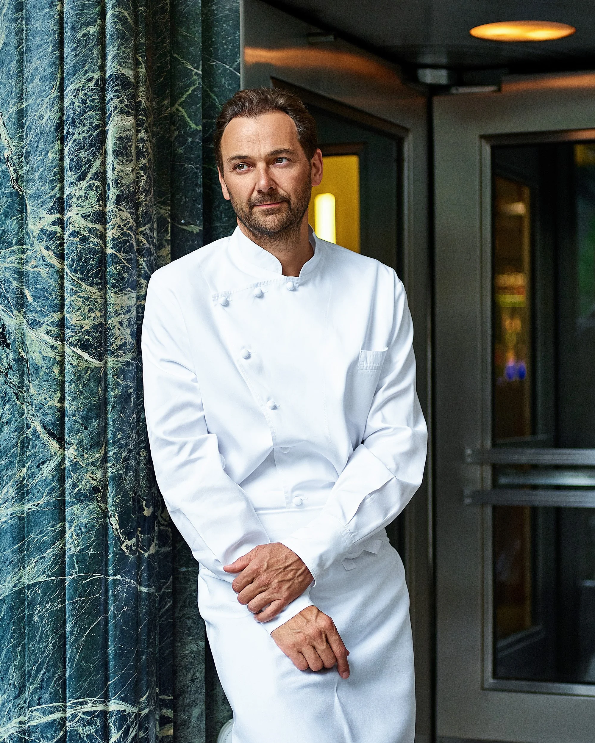 A male chef in a white uniform standing inside a modern restaurant or hotel, with green marble behind him and a dark ceiling with warm lighting.