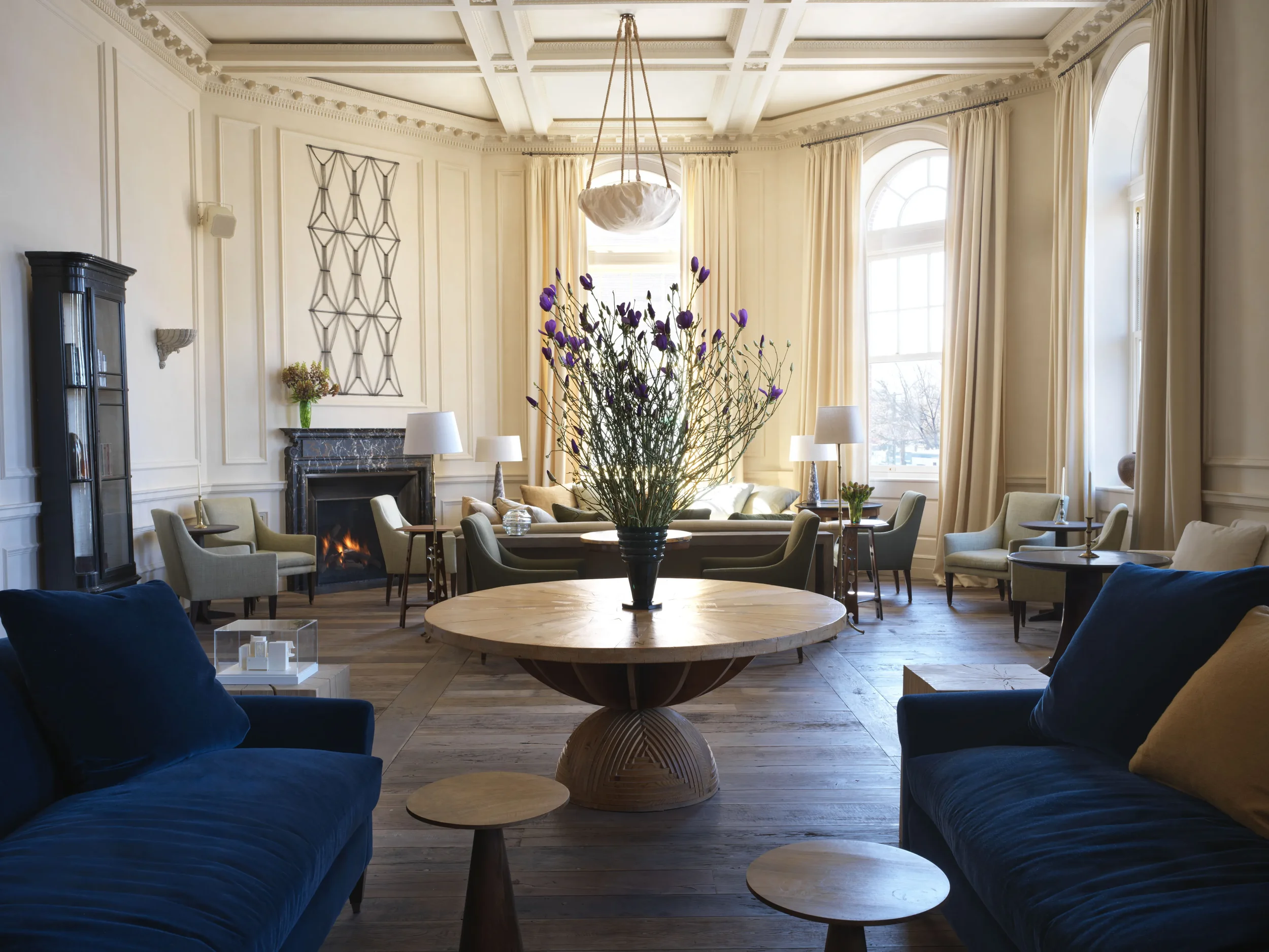 A spacious, well-lit living room featuring cream-colored walls with decorative molding and large windows with beige curtains. The room has multiple armchairs and sofas arranged around a large round wooden coffee table, with a tall flower arrangement with purple flowers in the center. is a black fireplace with a marble mantel, adorned with a small vase with flowers. There is a dark glass display cabinet on the left side of the room and a ceiling light fixture hanging above the coffee table.