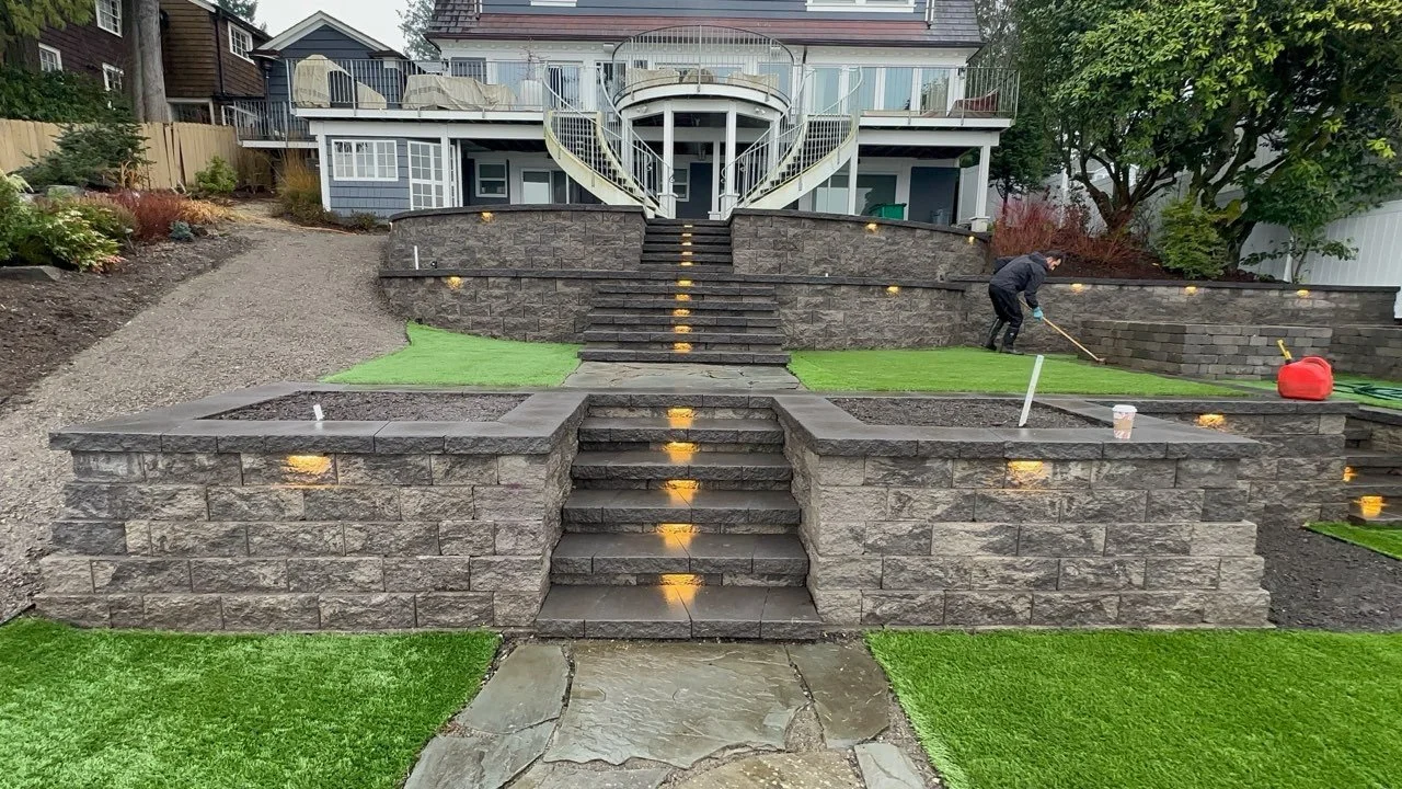 Landscape view of a backyard terraced with stone steps, retaining walls, and layered layouts, leading up to a house with a balcony and outdoor furniture. A person is working on the right side, preparing the area, with tools and supplies visible.