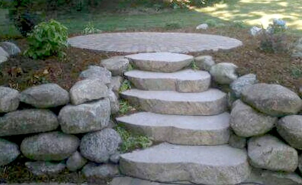 Stone staircase with multiple steps, flanked by stacked rocks on both sides, leading to a flat stone landing in a garden area.