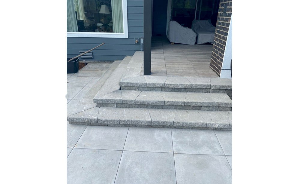 Outdoor patio with stone steps leading up to a covered porch area with a sliding glass door, gray siding, and a brick wall.