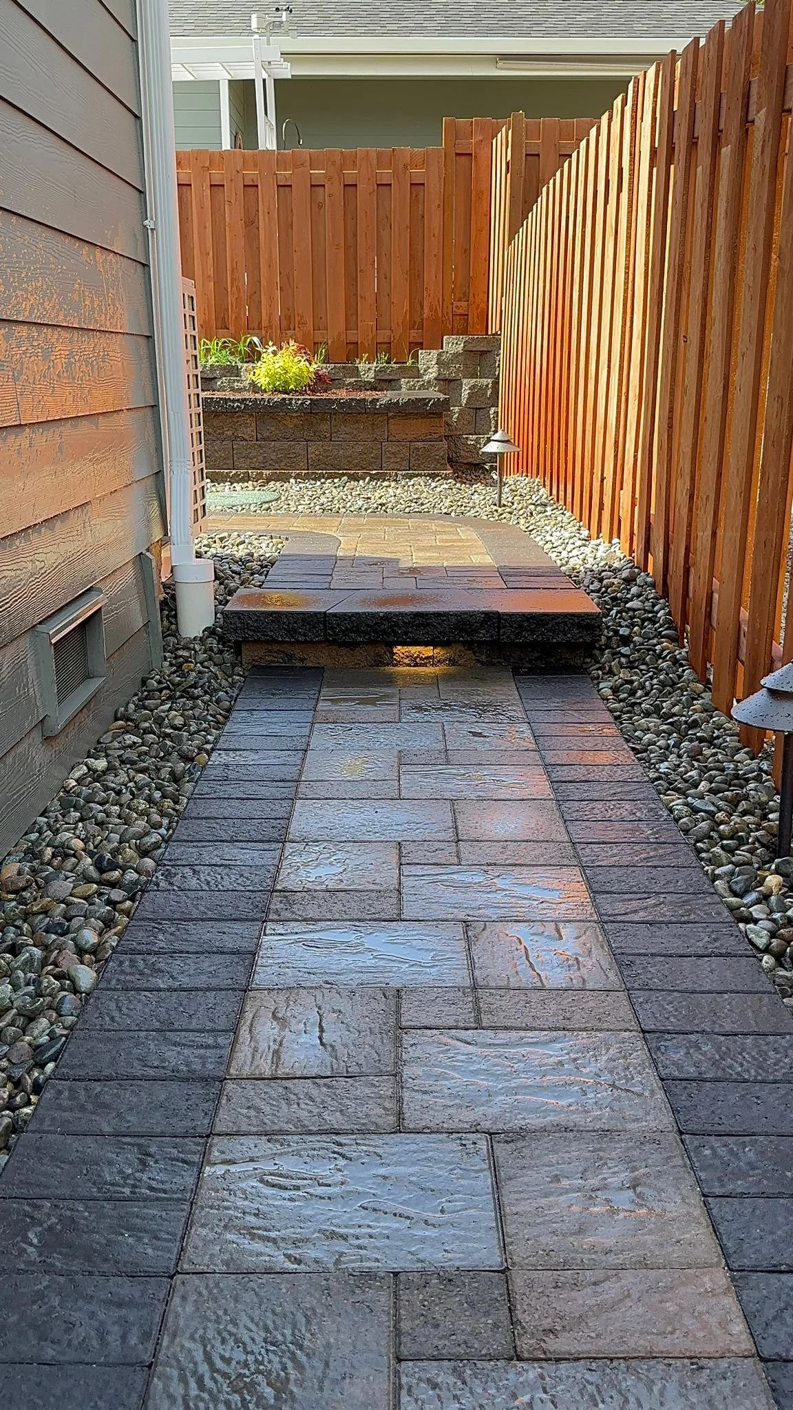 Clean wet paved garden path with stone borders, wooden fence on the right, and house wall on the left, with small landscaped area at the end of the path.