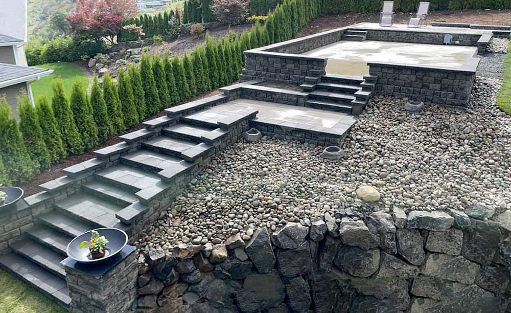 Backyard with a multi-level stone staircase, a gravel area, a stone retaining wall, and a small pool or hot tub with a concrete deck, surrounded by shrubs and trees.