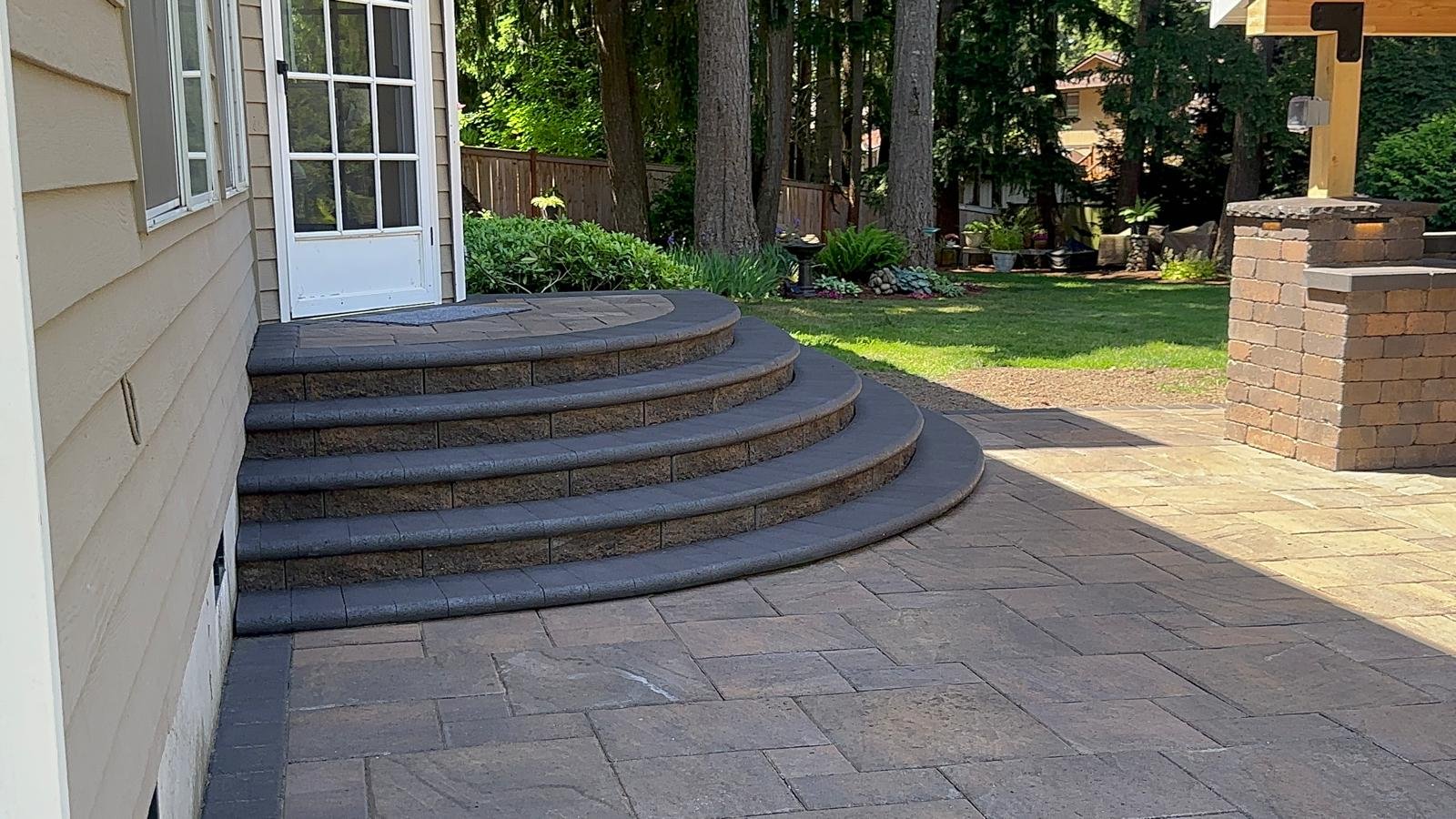 Backyard patio with stone steps and pavers, garden beds with plants, trees, and a wooden fence.