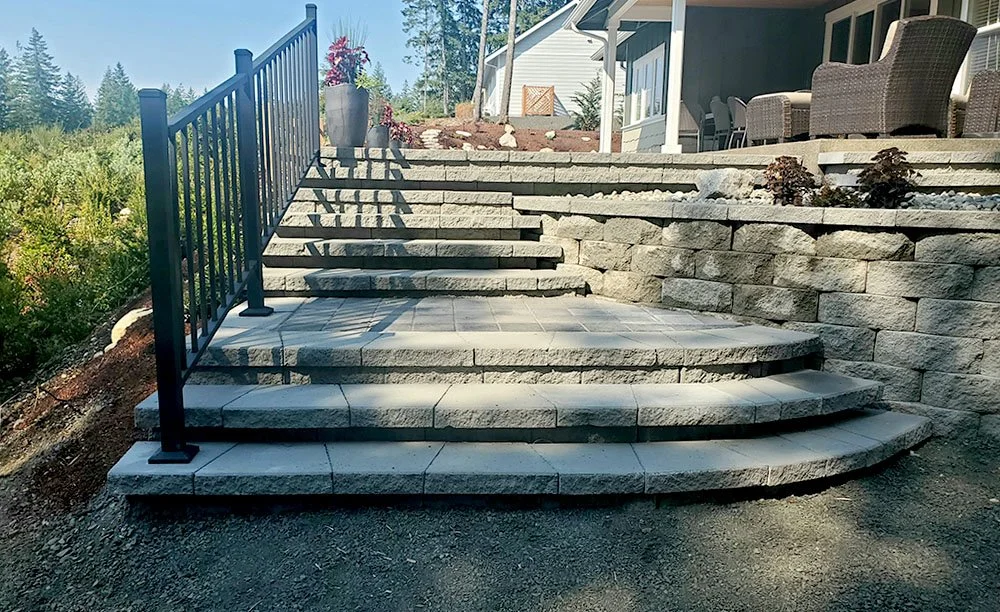 Outdoor stone staircase leading to a patio with outdoor furniture, railing on the left, and plants on the right, next to a house with a porch.