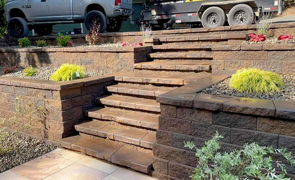 Stone staircase leading to a raised garden bed with plants and flowers in a landscaped yard, with vehicles in the background.