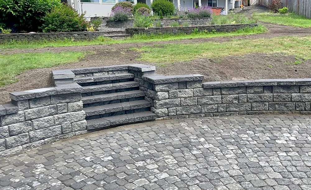 Stone steps with a small retaining wall leading to a grassy garden area with plants and flowers, in front of a house.