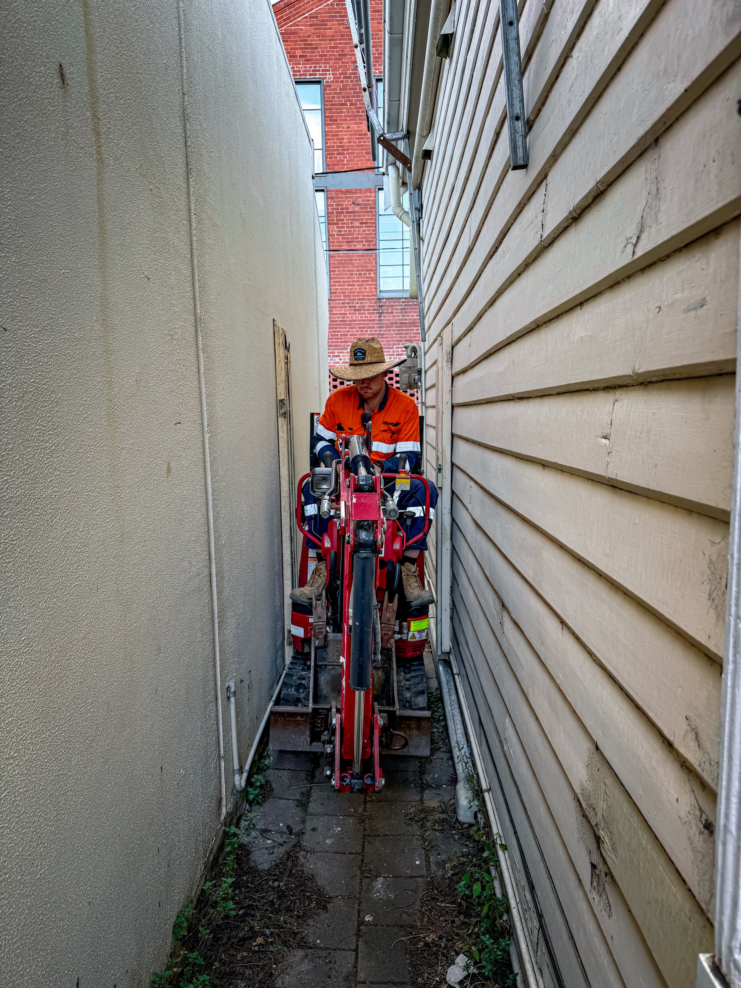 Worker operating a mini excavator in a tight access, narrow alley between a beige stucco building and a yellow wooden house.