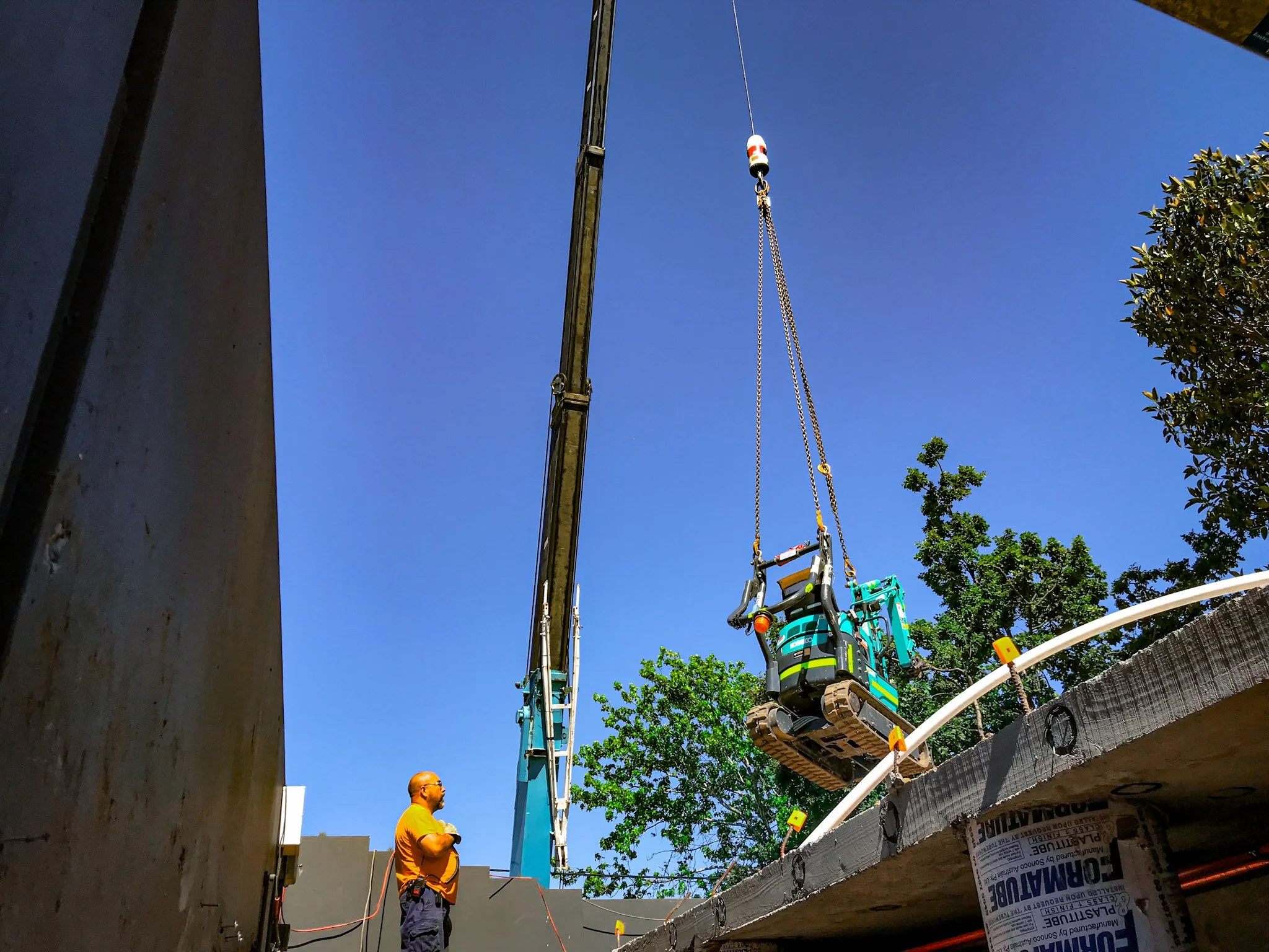 Construction worker observing a crane lifting a small excavator over a wall a construction site with trees in the background during daytime.