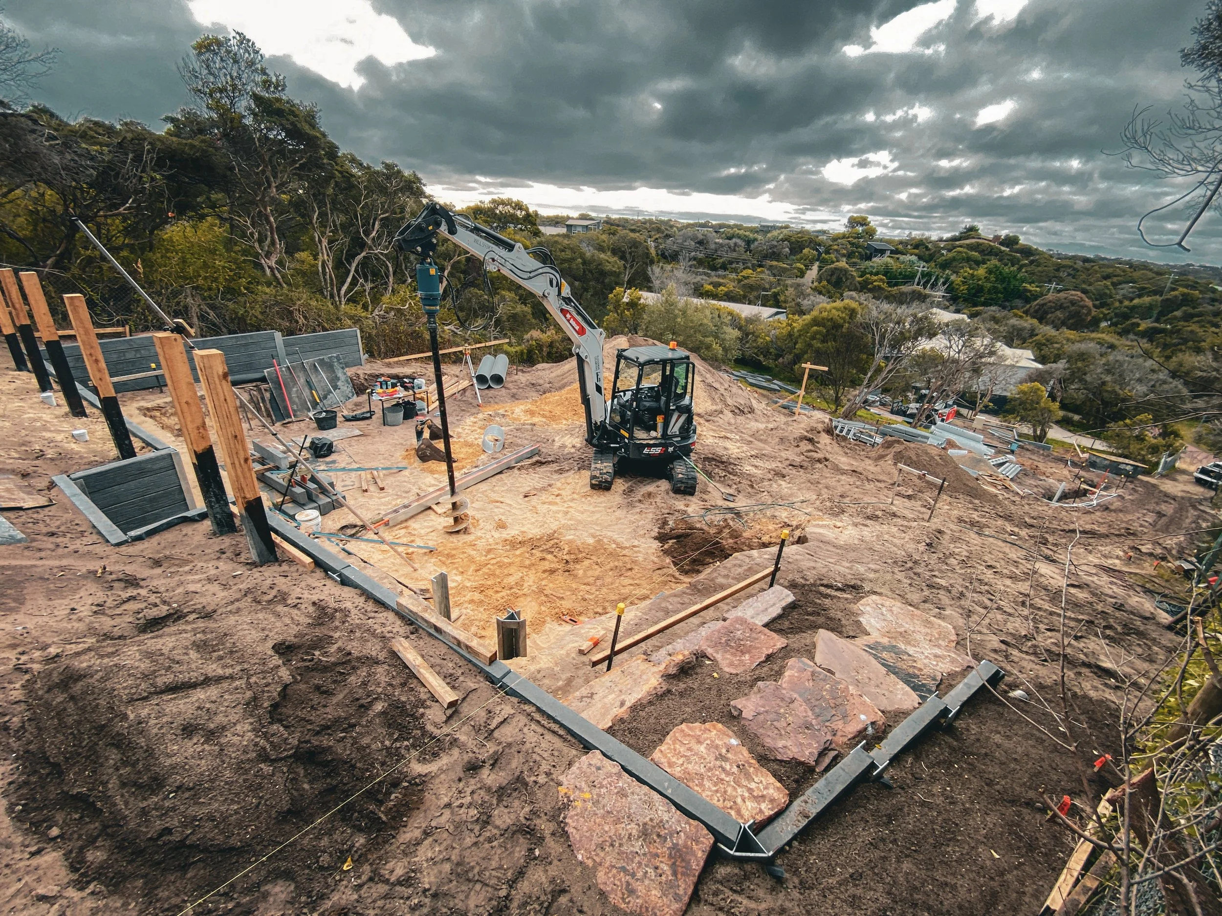 Construction site on a steep sandy hillside with a small excavator and auger, tools, and materials, rock stairs, in the process of constructing retaining walls, under cloudy sky.