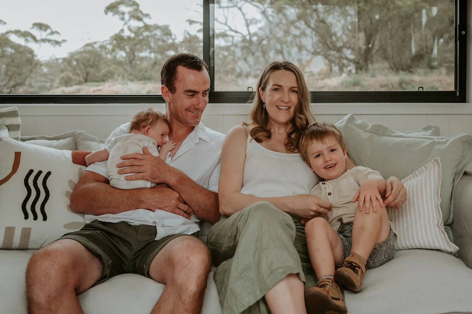 A family sitting on a sofa by a window with two children, one an infant held by the father and the other a toddler sitting with the mother, in a cozy living room.